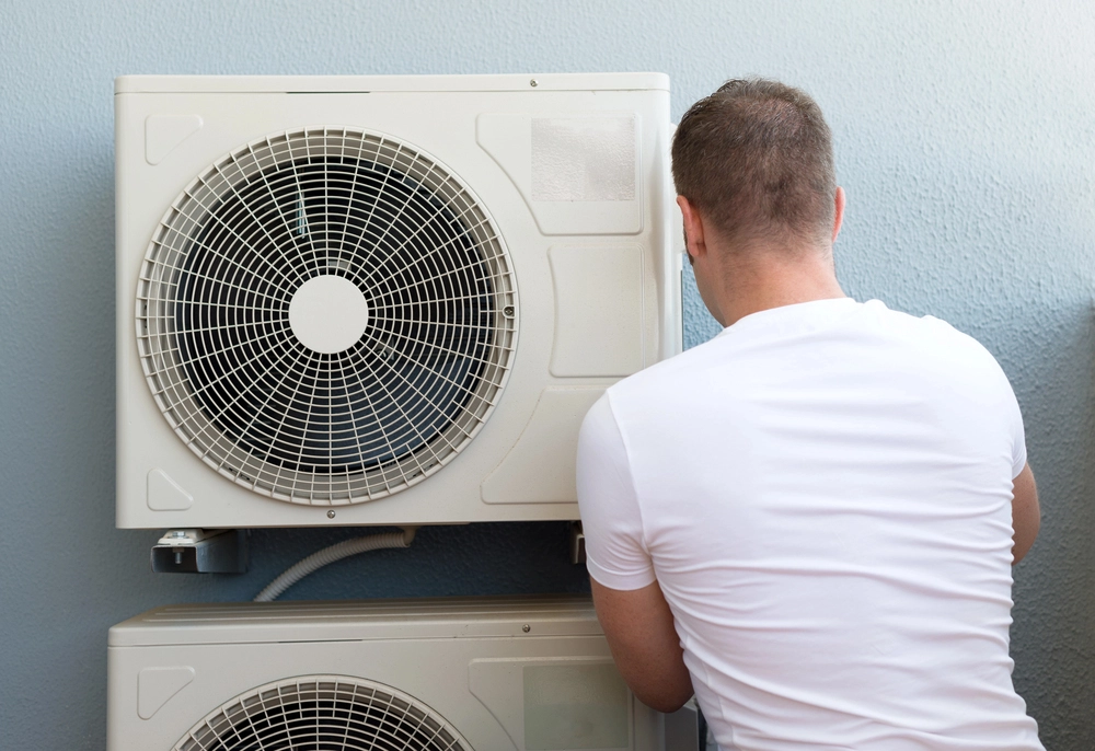 Technician servicing an outdoor HVAC condenser unit mounted on a wall.