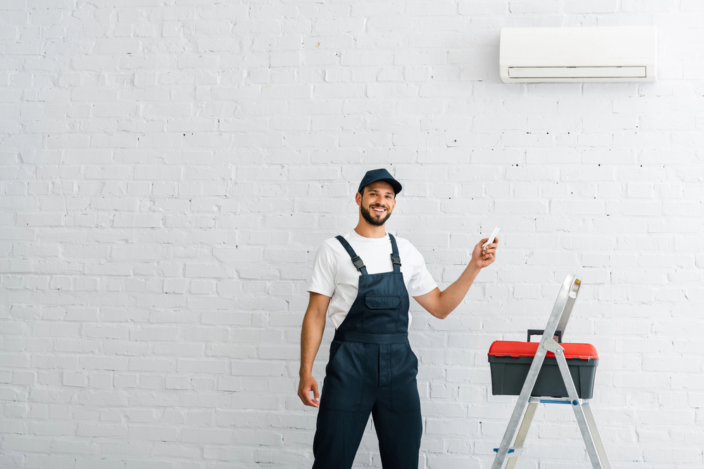 HVAC installer smiling while standing next to a wall-mounted air conditioner and tool kit.
